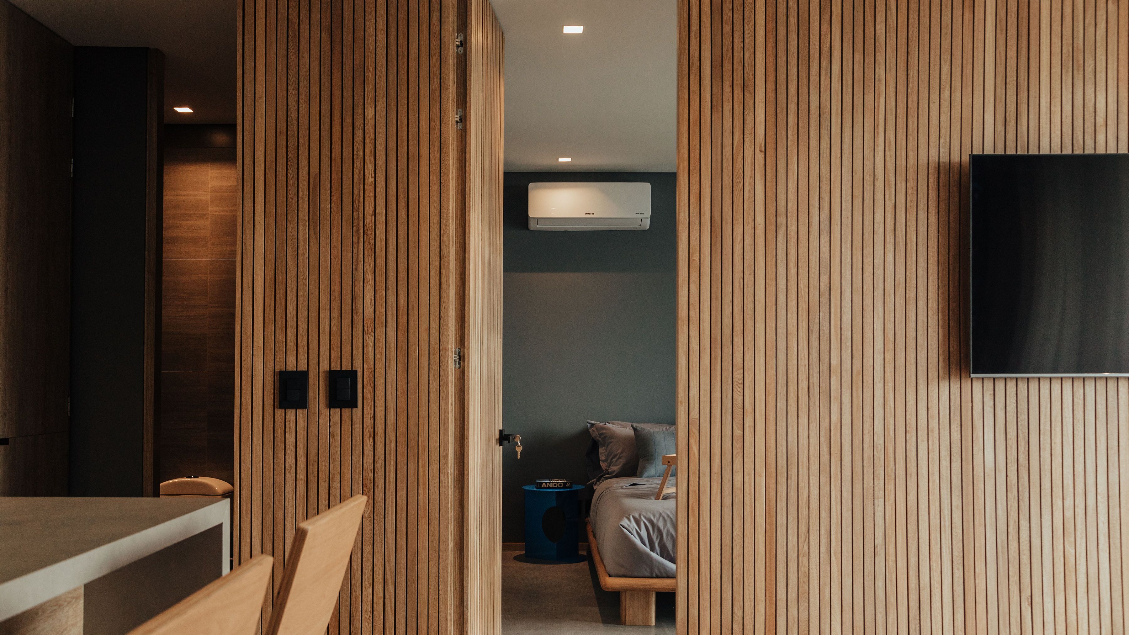Une chambre moderne avec un lambris en bois, un lit, une table de chevet, une petite table d’appoint bleue et un téléviseur fixé au mur.