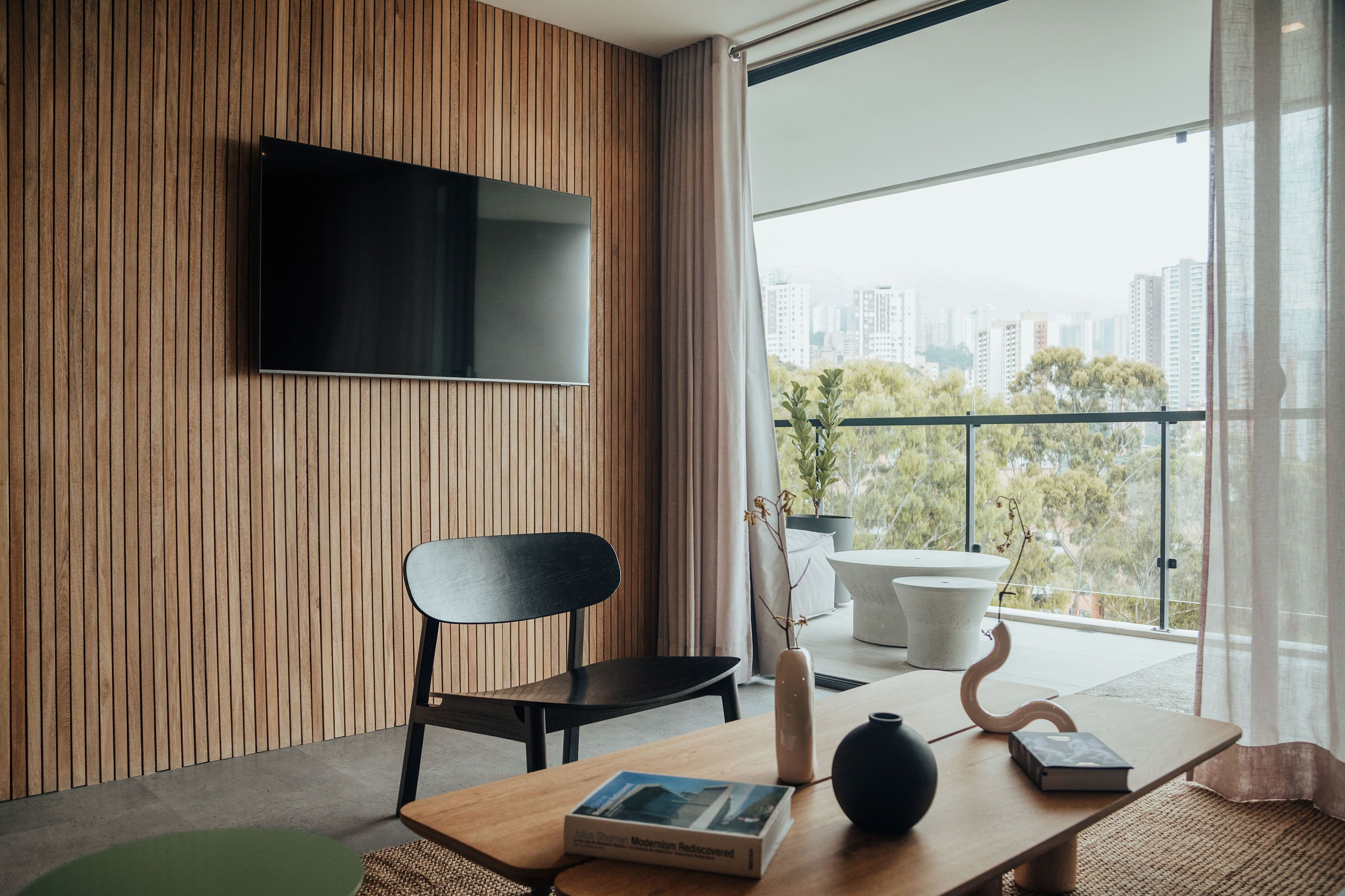 Un salon moderne avec un mur recouvert de panneaux de bois, un téléviseur à écran plat, un fauteuil noir, une table en bois avec un livre, un vase et une plante en pot.