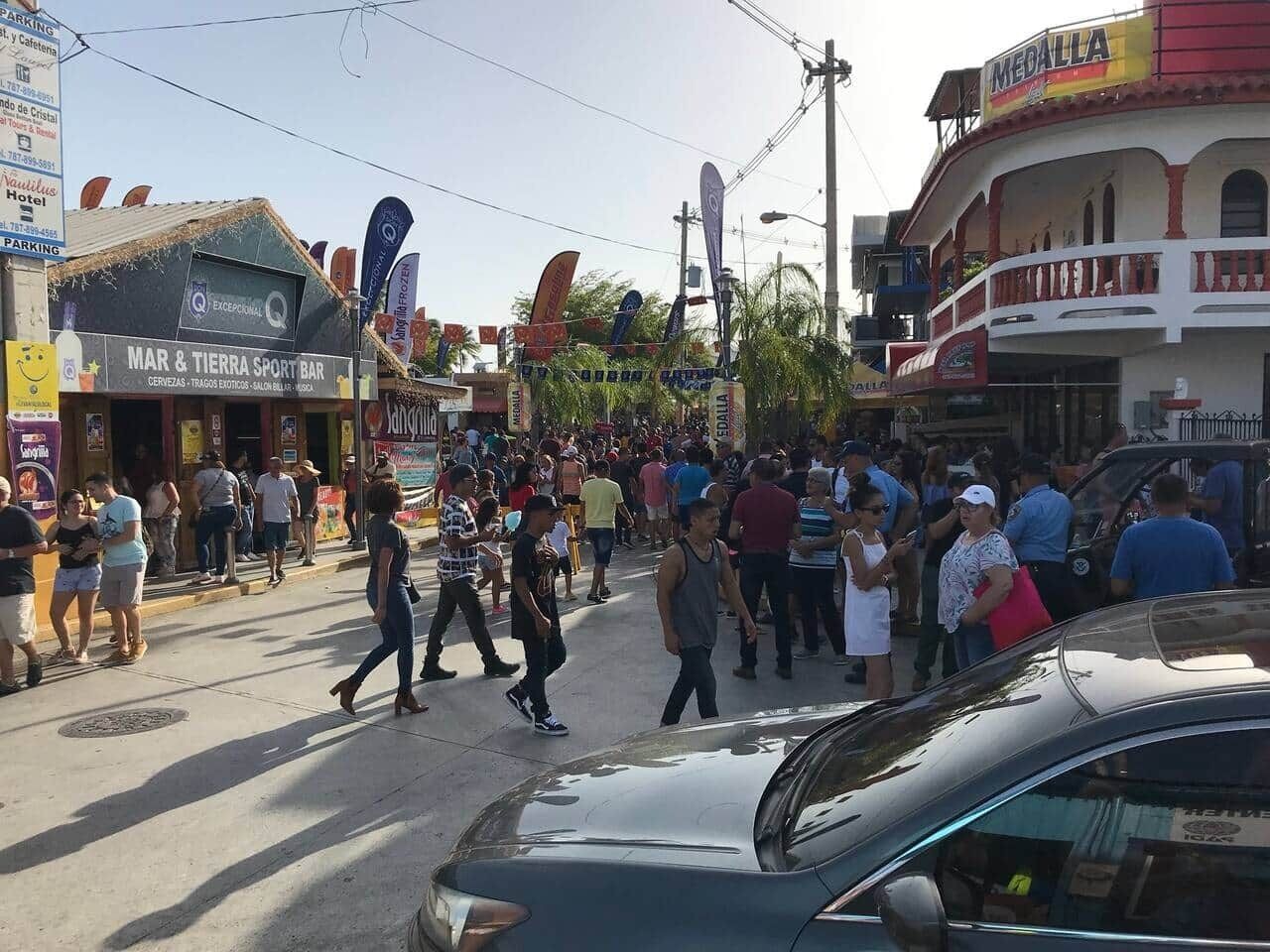 Rua movimentada com pedestres e carros, apresentando um bar esportivo 'MAR & TIERRA SPORT BAR' e um edifício 'MEDALLA'.
