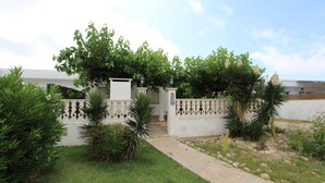 Una propiedad residencial con entrada pavimentada, un muro blanco con ornamentación de hierro forjado y abundante vegetación, incluyendo árboles y arbustos.