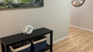 Ein Zimmer mit einer schwarzen Konsolentisch, zwei Stoff-Aufbewahrungsbehältern, einem weißen Dekorationsobjekt, einem wandmontierten Spiegel und einem Gemälde von einer Küstenlandschaft.
