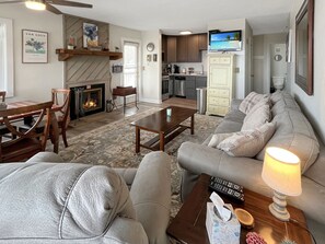 A living room with a fireplace, sofa, coffee table, and a television mounted above the kitchen.