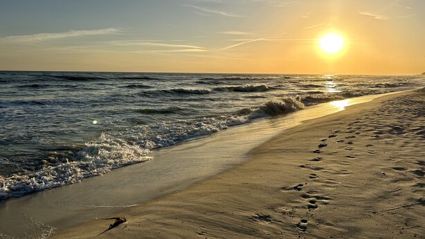 En strand med fodspor der fører ned til vandet, bølger og en solnedgang.