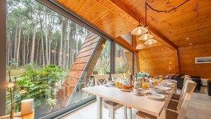 A modern cabin with a dining area featuring a long table set for breakfast, large windows offering a view of a forest, and wooden interior design. - Woodpecker, Weybourne, Norfolk (Weybourne)