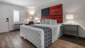 A bedroom with a large bed, a wall-mounted American flag, two bedside tables with lamps, a chair, and a window with blinds. - Super 8 by Wyndham Daleville/Fort Novosel (Daleville)