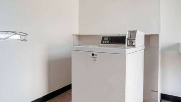 A compact, white laundry machine in a hallway with plain white walls and a black baseboard.