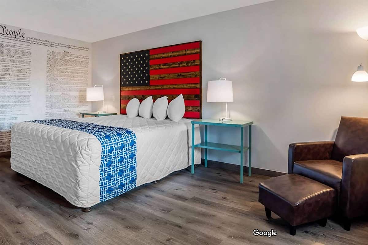A hotel room with a large bed, a teal nightstand, a brown leather armchair, and a wall with a large American flag and a wall of text.