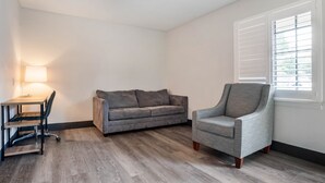 A room with a grey sofa, a grey armchair, a wooden desk with a lamp, and a window with blinds. - Super 8 by Wyndham Daleville/Fort Novosel (Daleville)