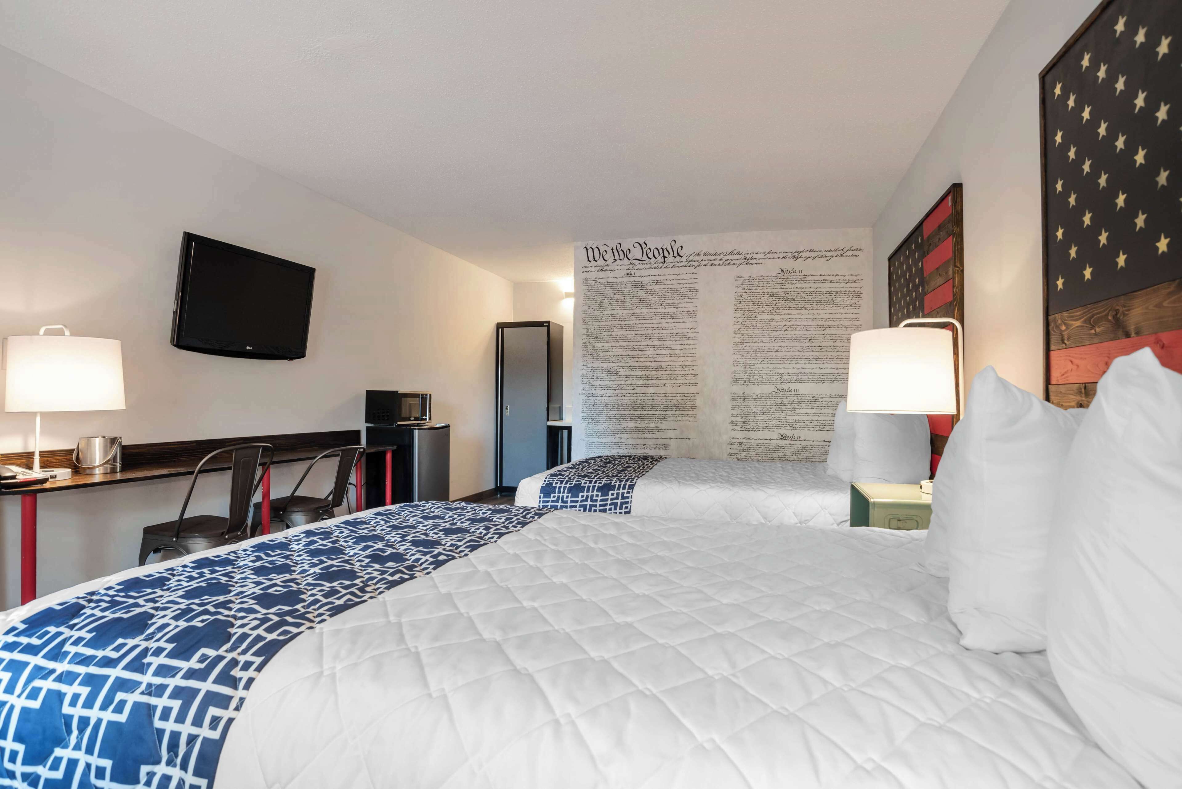 A hotel room with two beds, a desk, a TV, and a wall with an American flag and a large text display.