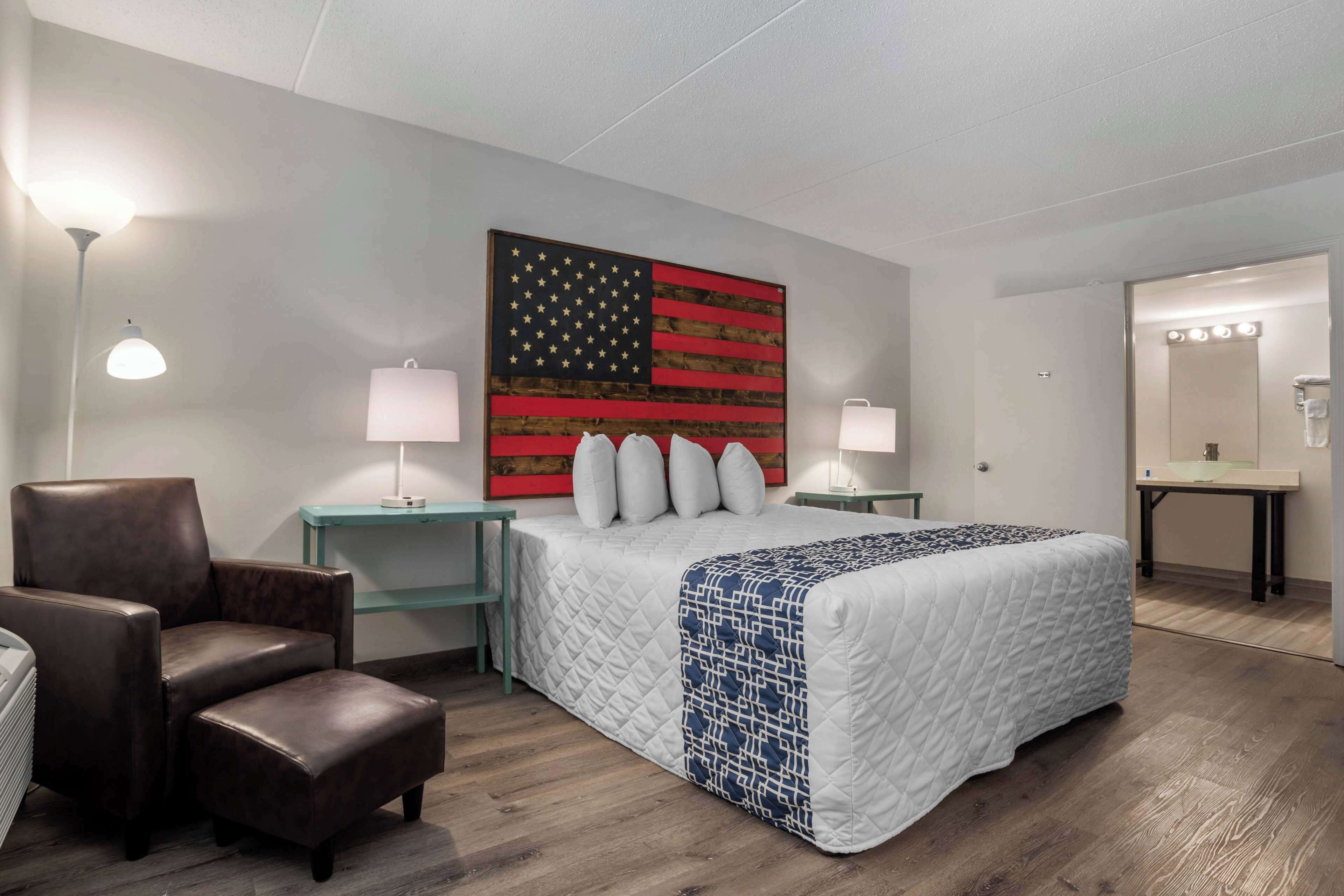 Habitación de hotel con un mural grande de la bandera estadounidense, una cama tamaño queen con colcha estampada, un sillón de cuero, dos mesitas de noche con lámparas y un tocador con espejo e iluminación.