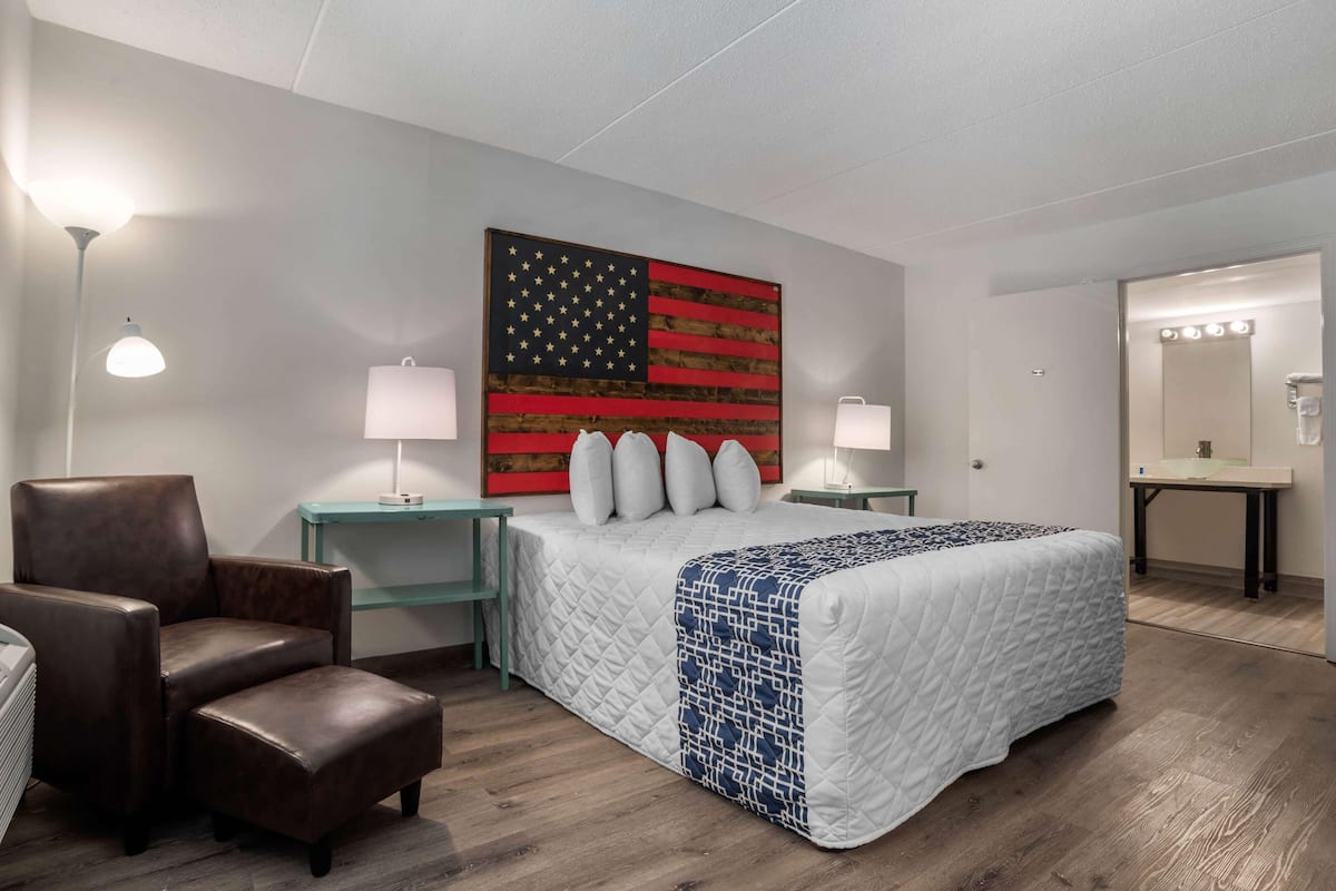 A hotel room with a bed, a chair, a nightstand, a lamp, a wall with a flag, and a bathroom with a mirror and lights.