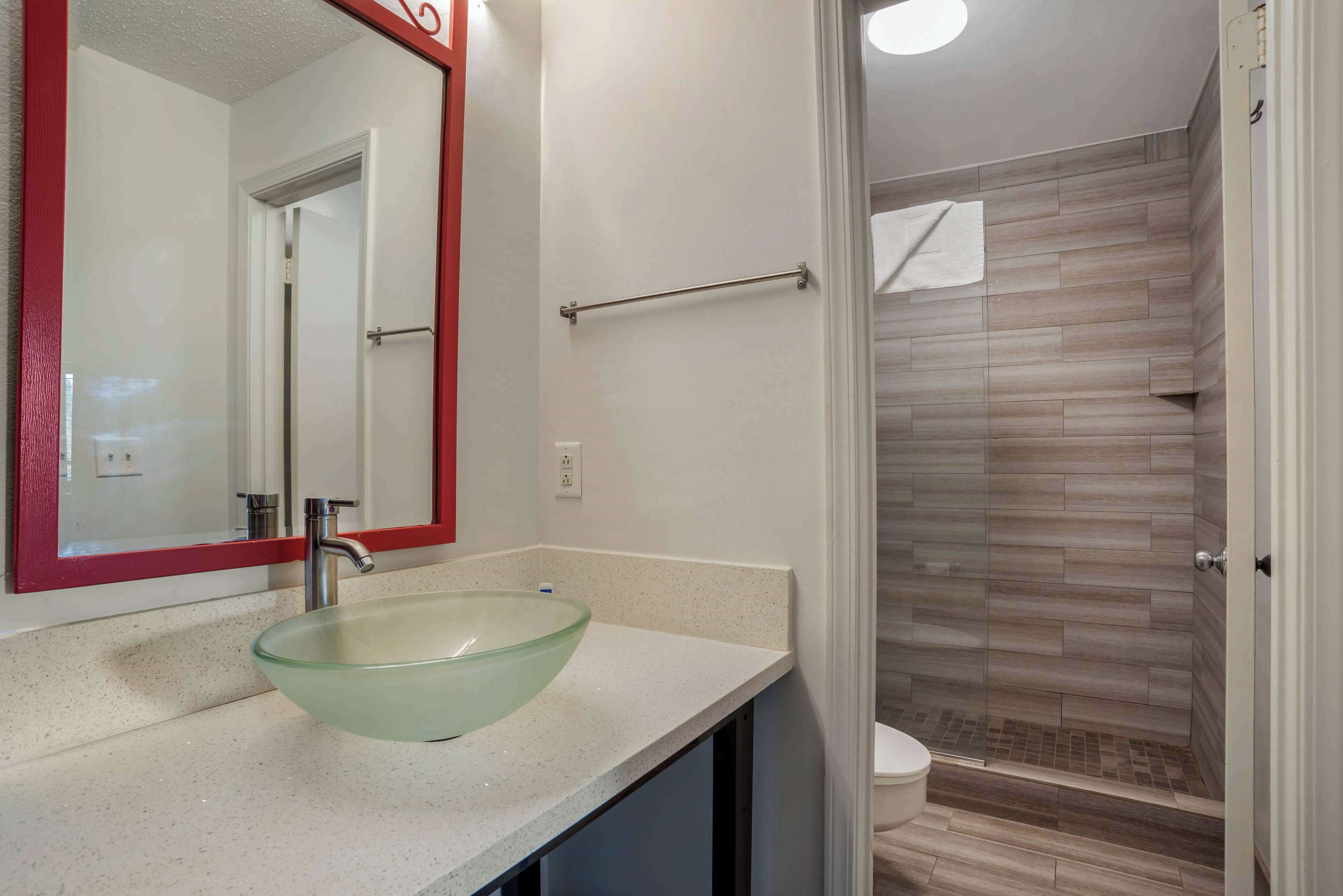 Un baño con un lavabo de vidrio, un espejo con marco rojo, un perchero y una ducha con azulejos de madera.