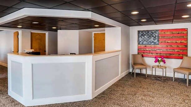A reception area with a white reception desk, two beige armchairs, a small table with flowers, and a large American flag painting on the wall. - Super 8 by Wyndham Daleville/Fort Novosel (Daleville)