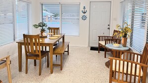 A dining area with a wooden table and chairs, a ceiling fan, and a view of the street through large windows. - Treasure Coast Pool Home - Walk to Downtown Jensen Beach. (Jensen Beach)