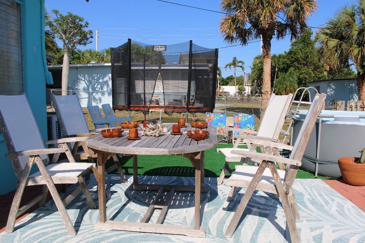Un patio exterior con una mesa de madera redonda, sillas y un trampolín bajo un toldo.