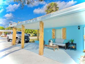 Un espacio exterior techado con pared azul, pilares de madera, una camioneta blanca y un sofá con cojines.