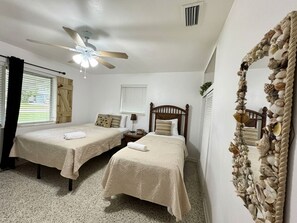 Une chambre avec deux lits simples, un ventilateur de plafond, un miroir orné d’une bordure en forme de coquillage et une fenêtre avec des stores.
