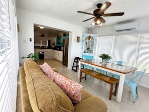 Un salon avec un canapé, une table à manger entourée de chaises, un ventilateur de plafond et, au fond, une cuisine.