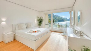 A modern bedroom with a bed, bedside tables, a dresser, a large window with a view of mountains and a lake, and a mirror on the dresser. - Honeymoon with Panoramic Lake-View, Castagnola (Lugano), Switzerland (Castagnola (Lugano))