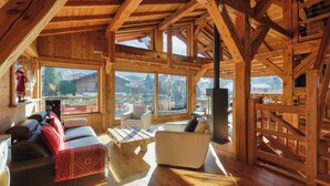 A spacious wooden interior with a sofa, armchairs, and a TV. Large windows offer views of the outdoors.