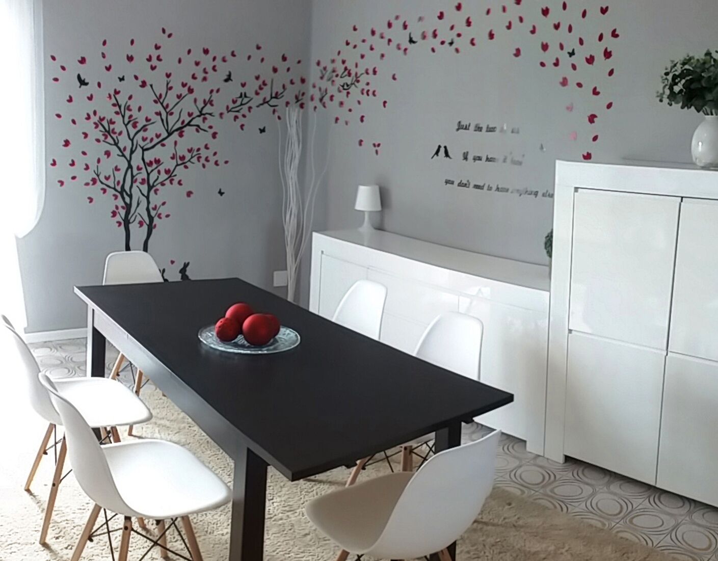 Uma área de refeições moderna com uma mesa preta, cadeiras brancas e uma parede decorativa com uma árvore e corações.