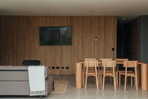 Uma sala de estar moderna com parede de painéis de madeira, uma TV de tela plana, um sofá, uma mesa de centro e uma área de jantar com cadeiras e mesa de madeira.