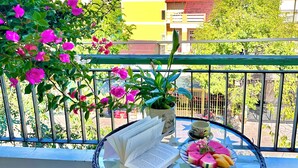 Une table ronde en verre, sur laquelle se trouvent un livre, une plante en pot et des fruits, sur un balcon.