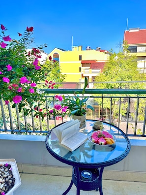 Una mesa redonda de vidrio con un libro y fruta, una planta en maceta y una barandilla con flores rosadas.