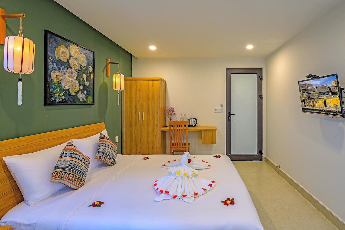 Quarto de hotel com cama, almofadas, guarda-roupa de madeira, escrivaninha com cadeira, televisão na parede e um quadro.