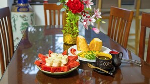 Une table à manger avec une assiette de pastèque et de bananes, un vase rempli de fleurs et une tasse de café.