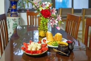 Una mesa de comedor con una fuente de sandía y plátanos, un florero con flores y una taza de café.