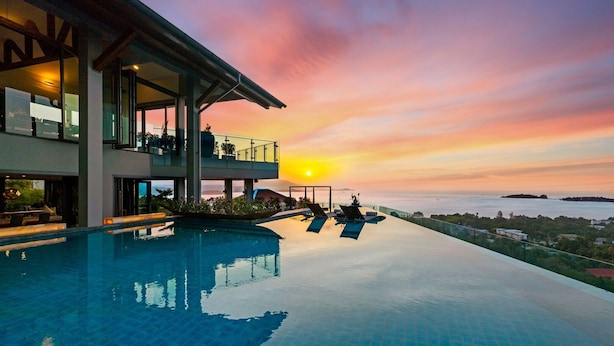 Ein moderner Infinity-Pool mit Sonnenuntergangsblick, ein großes Haus mit Außenbereich und einem klaren Himmel.