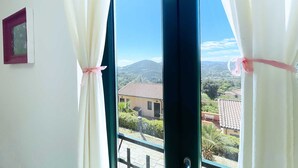 A window with white curtains tied with pink ribbons, offering a view of a hillside with buildings.