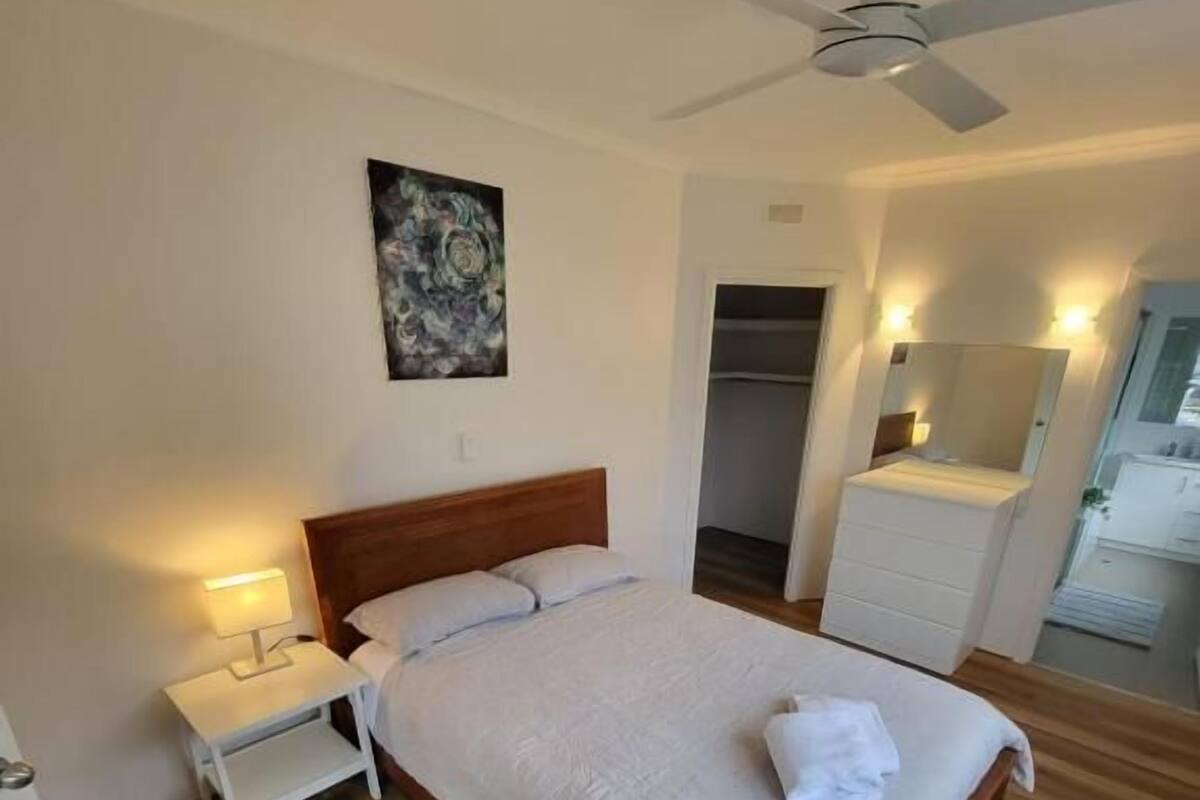 A neatly arranged bedroom with a wooden bed, a nightstand with a lamp, a ceiling fan, a painting on the wall, and a walk-in closet.