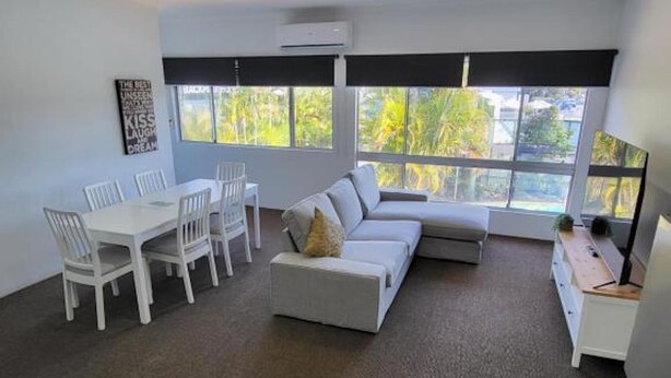 A modern living room with a sectional sofa, a dining table with chairs, and large windows offering a view of greenery outside. - Central Motel Mooloolaba Apartments   (Mooloolaba)