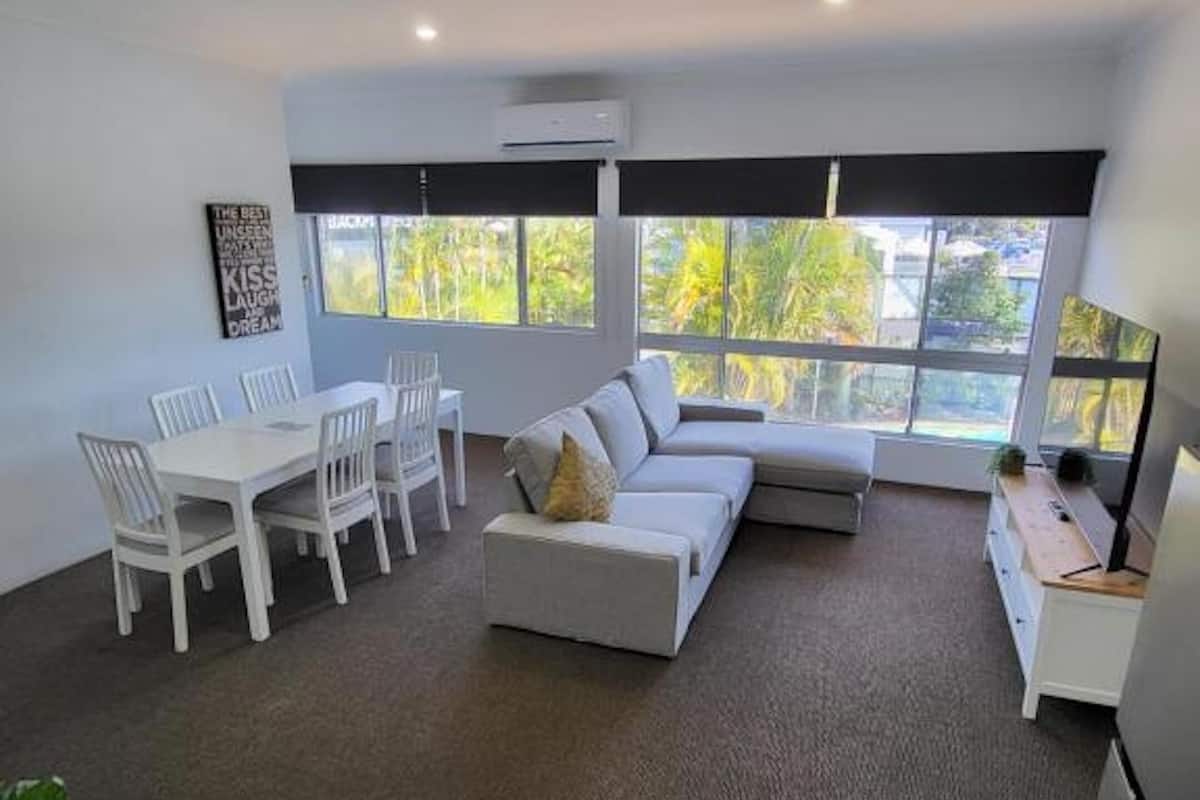 A modern living room with a sectional sofa, a dining table with chairs, and large windows offering a view of greenery outside.