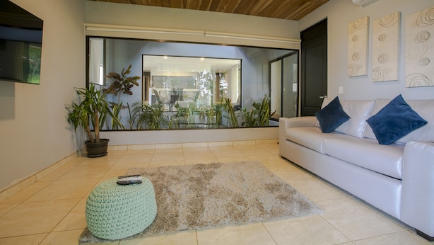 A modern living room with a white sofa, blue cushions, a green ottoman, and a large window overlooking a garden. - River Paradise Villa (La Fortuna)