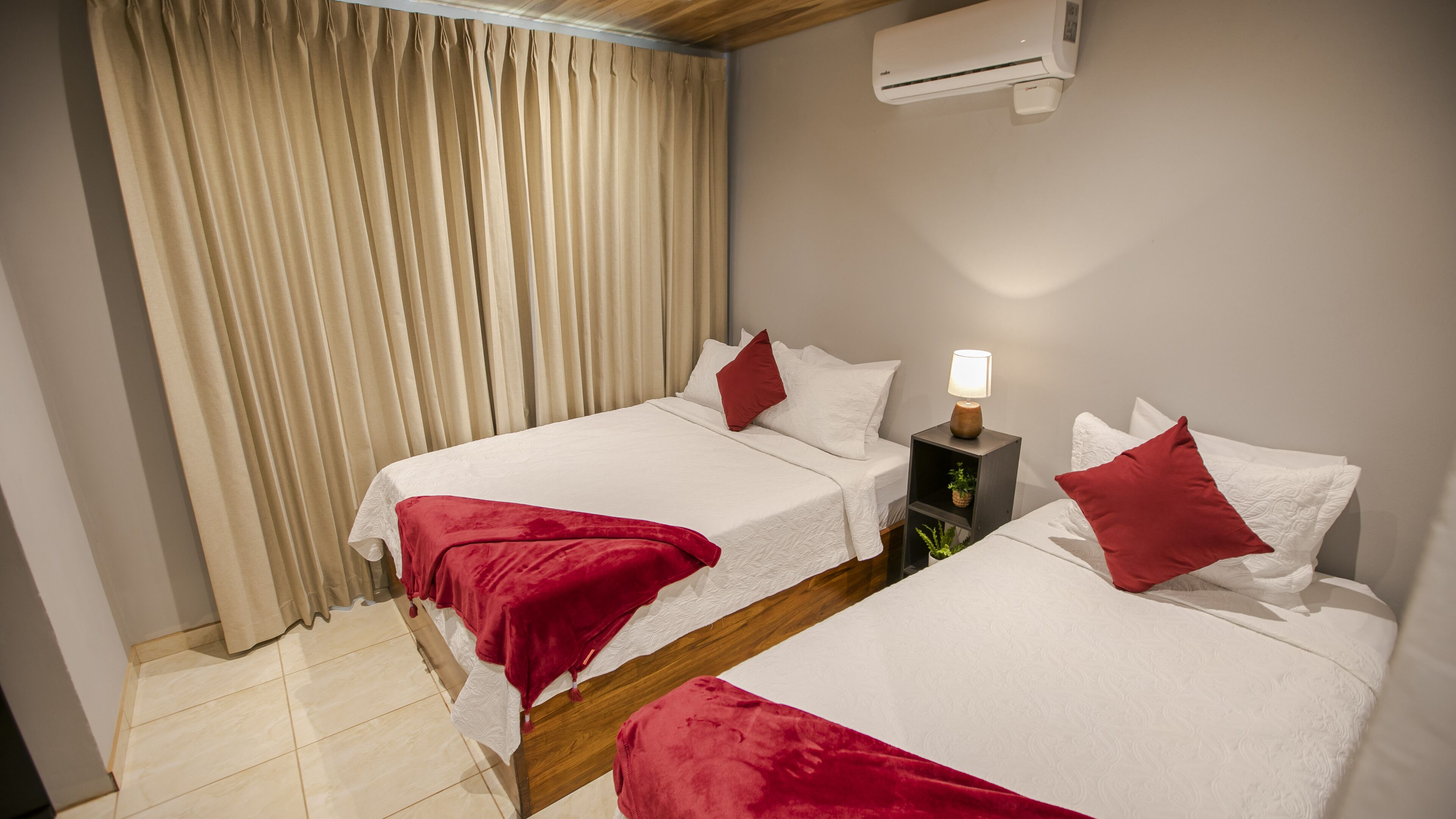 A hotel room with two beds, beige curtains, an air conditioning unit, a bedside lamp, and a small plant on a nightstand.