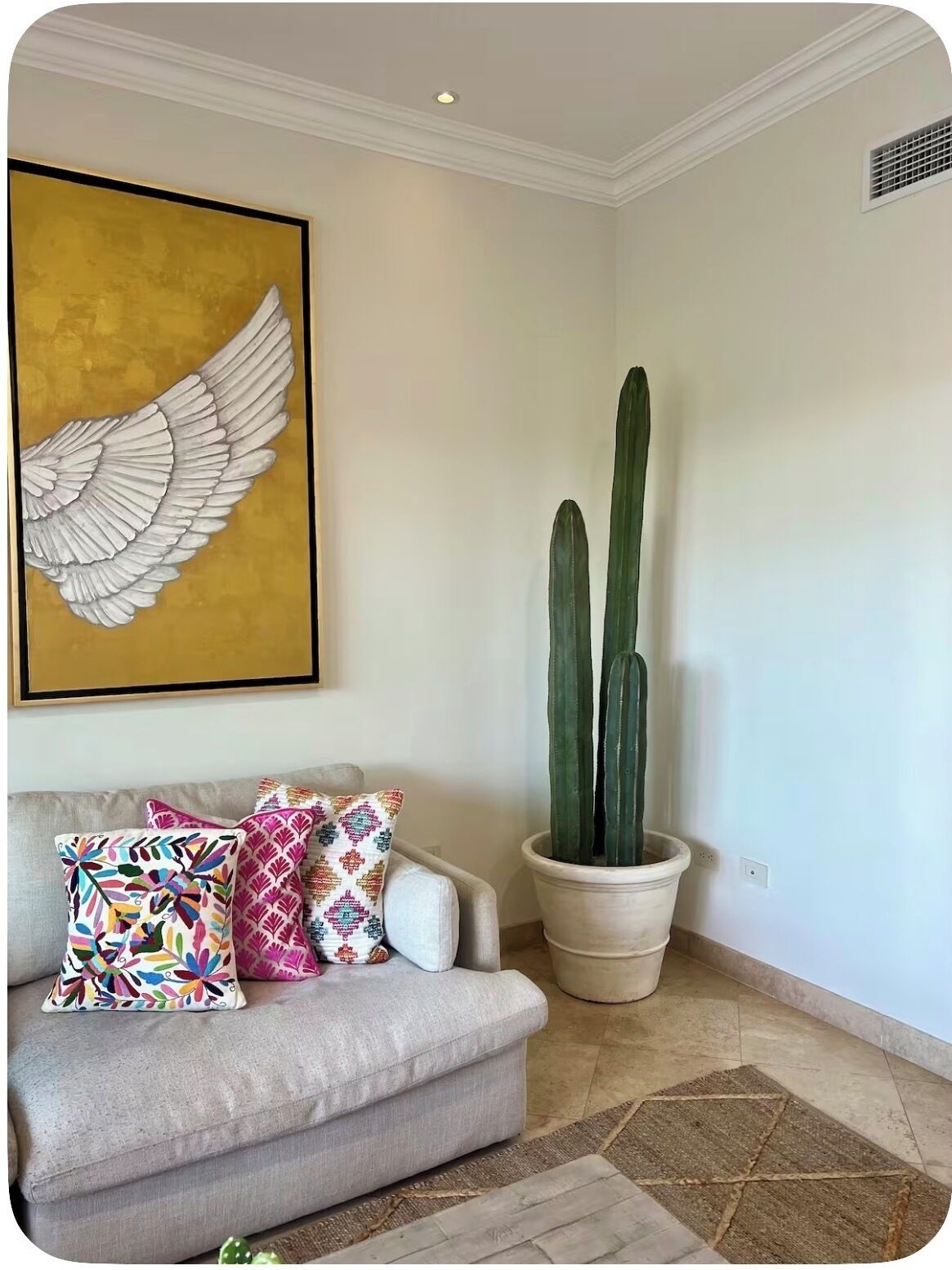 Un salon avec un canapé, des coussins colorés, une grande plante en forme de cactus et une œuvre d’art représentant des ailes, accrochée au mur.