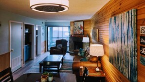 A cozy living room with a fireplace, wooden walls, a dining area with a table and chairs, and a large window.