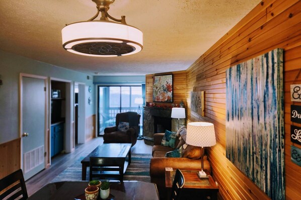 A cozy living room with a fireplace, wooden walls, a dining area with a table and chairs, and a large window.
