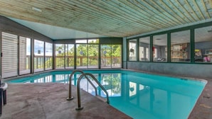 An indoor swimming pool with a concrete floor, metal handrails, and large windows offering views of the outside.