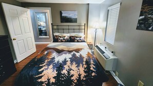 Un dormitorio con cama, mesita de noche, lámpara, ventana con persianas, ventilador de techo y puerta que da a una cocina.