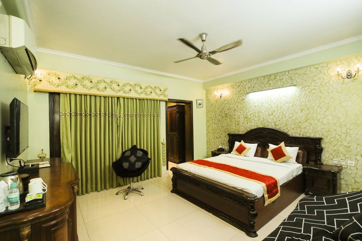 A hotel room with a bed, a desk with a chair, a ceiling fan, and a wall-mounted TV.