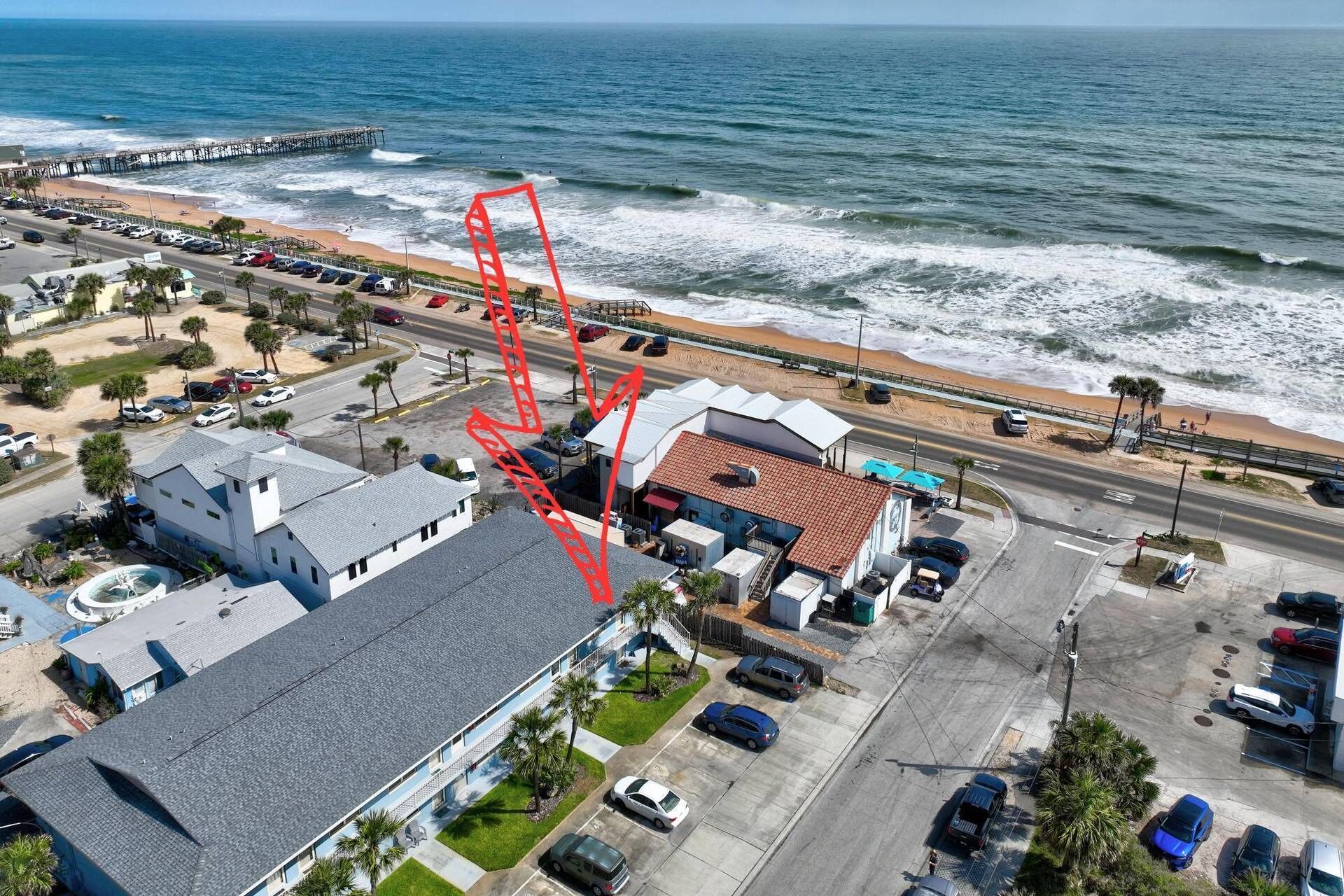 An aerial view of a coastal area with a beach, a pier, and a parking lot.