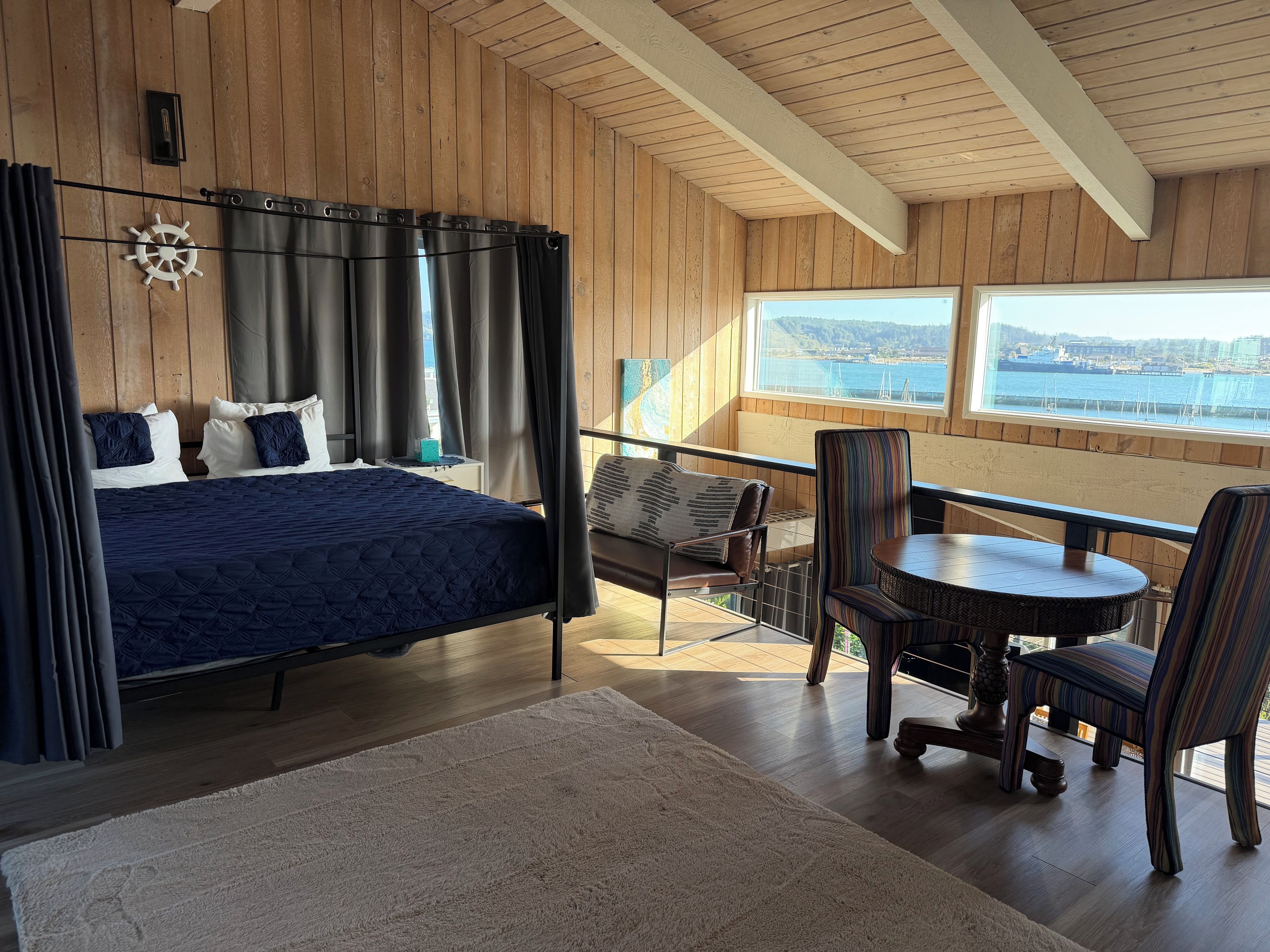 Een slaapkamer met een groot bed, uitzicht op zee, een balkon met een tafel en stoelen, en houten wanden.