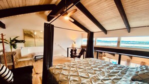 Un dormitorio con una cama grande, un escritorio, una silla y vistas de la ciudad a través de amplias ventanas.