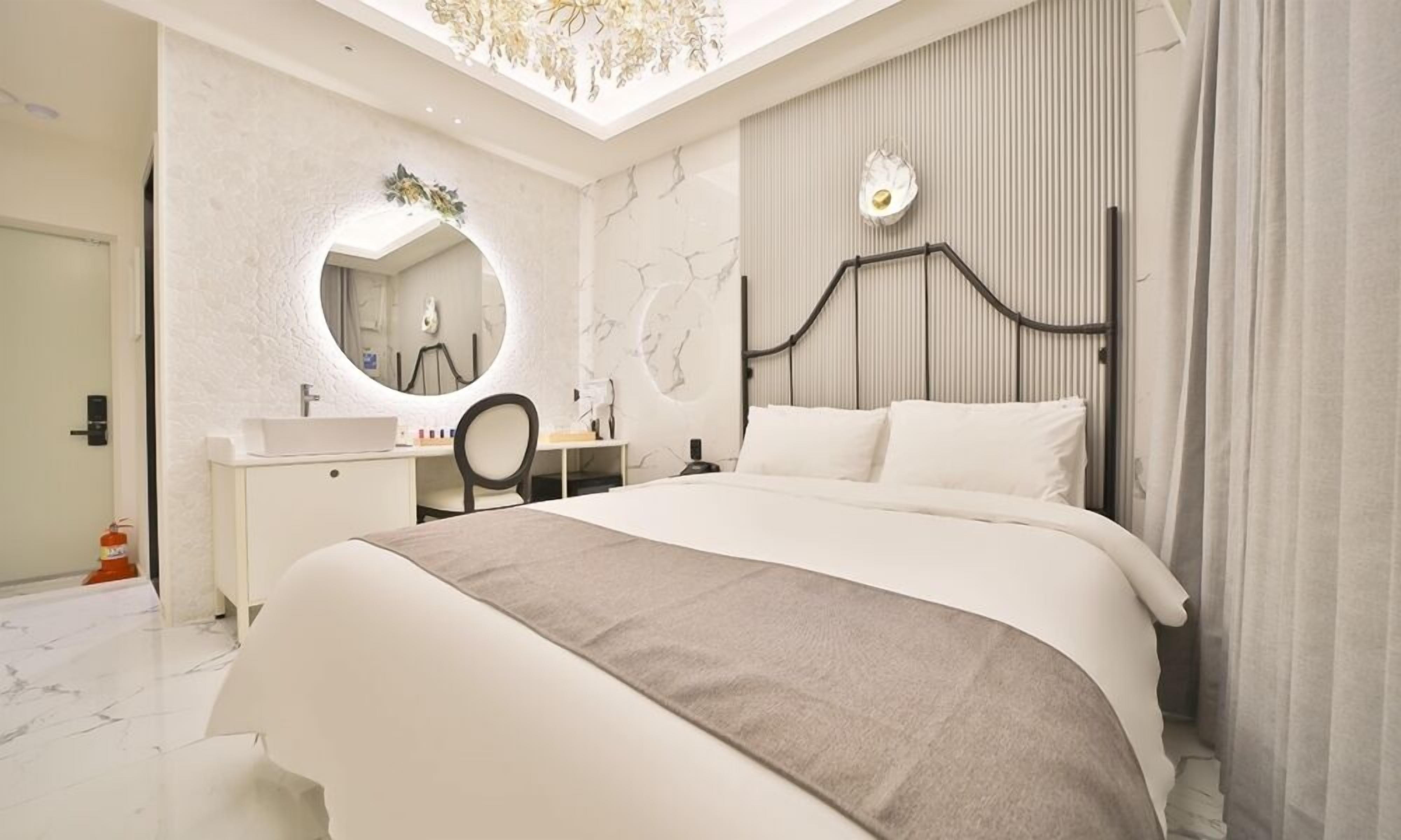 A modern bedroom with a large bed, a round mirror, a desk, and a bathroom visible through an open door.