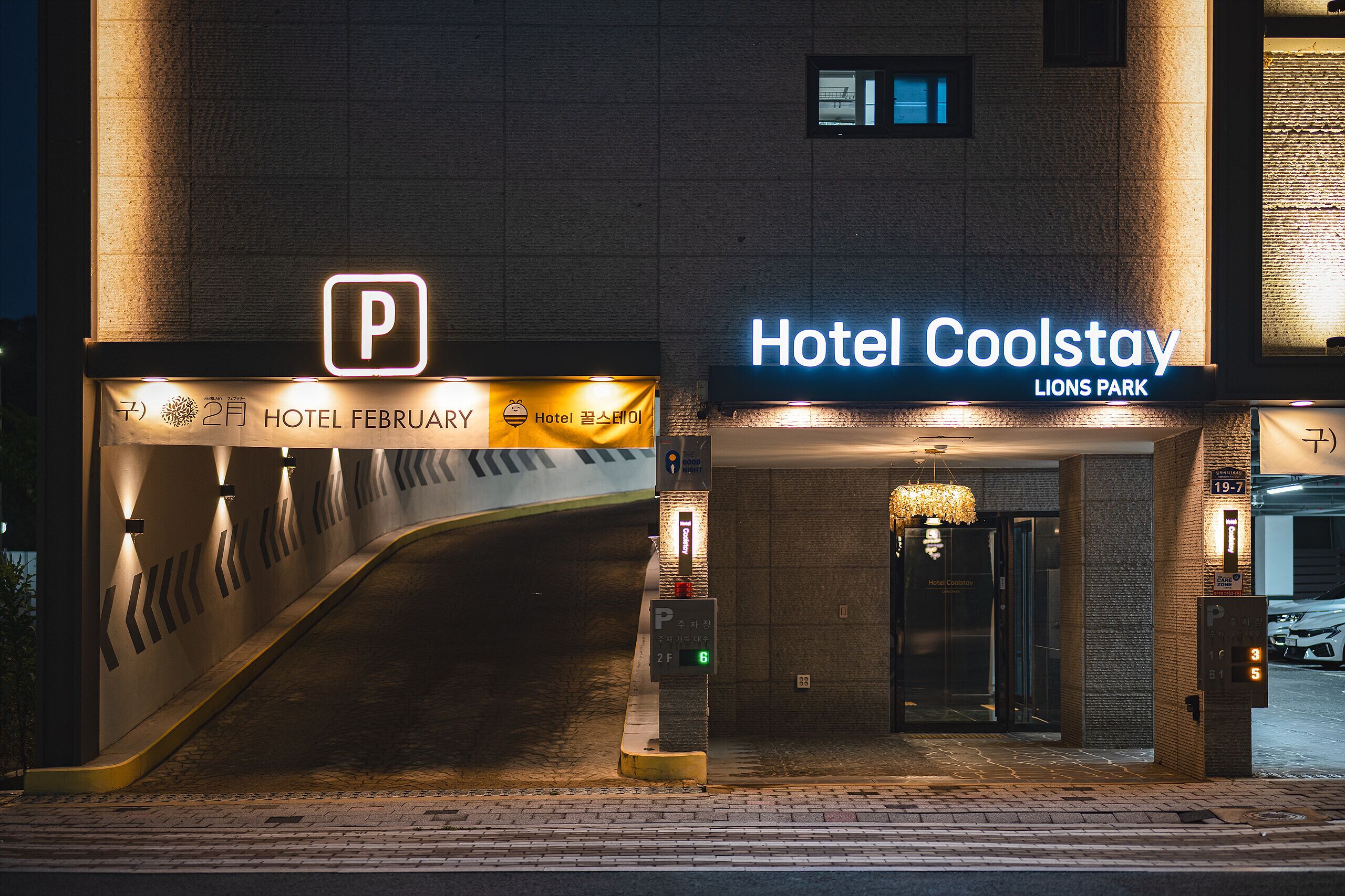 A hotel entrance with a parking sign, a neon sign for Hotel Coolstay, and a digital parking meter.