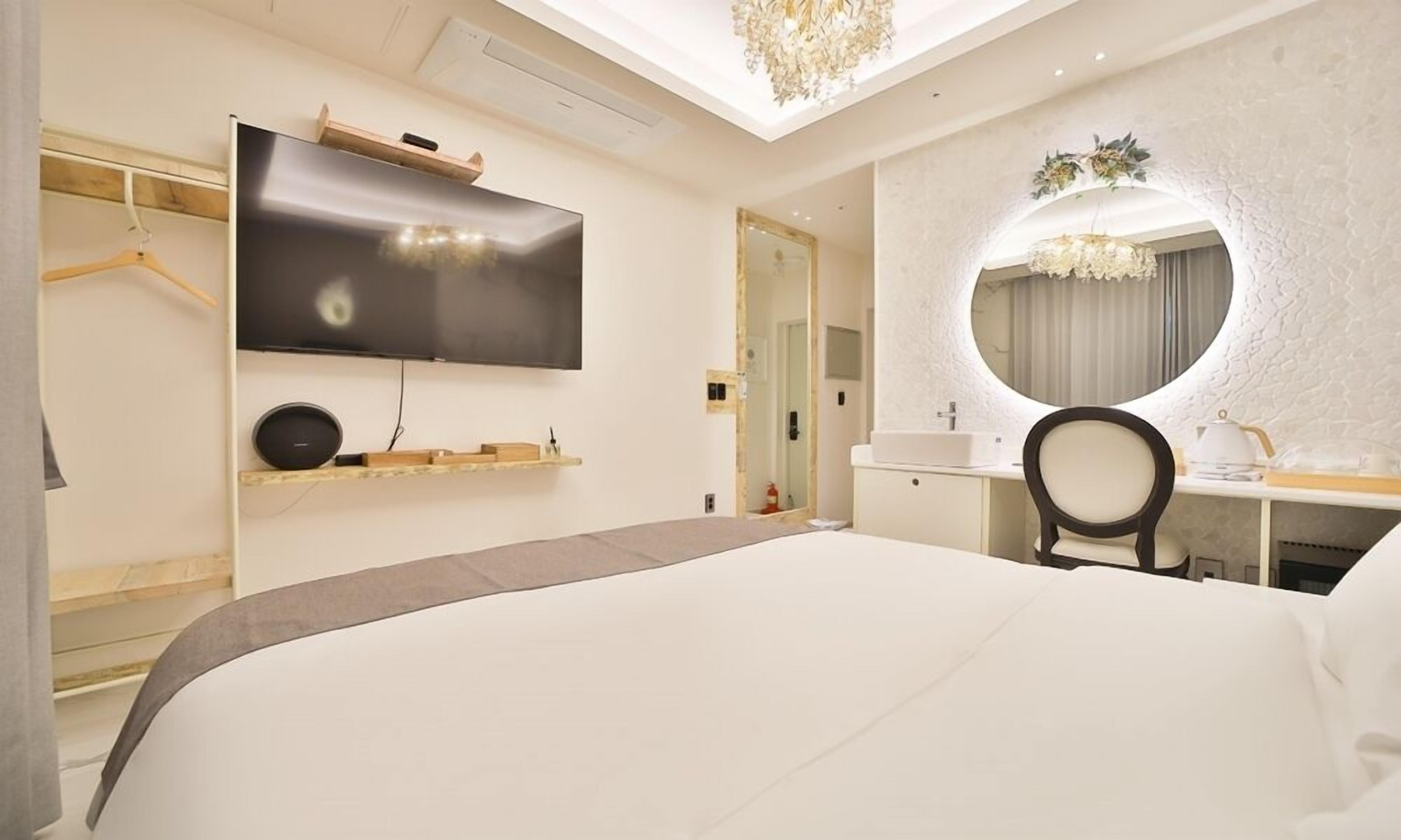 Une chambre d’hôtel moderne avec un grand lit, un téléviseur à écran plat fixé au mur, un miroir rond au-dessus du bureau et un lustre.